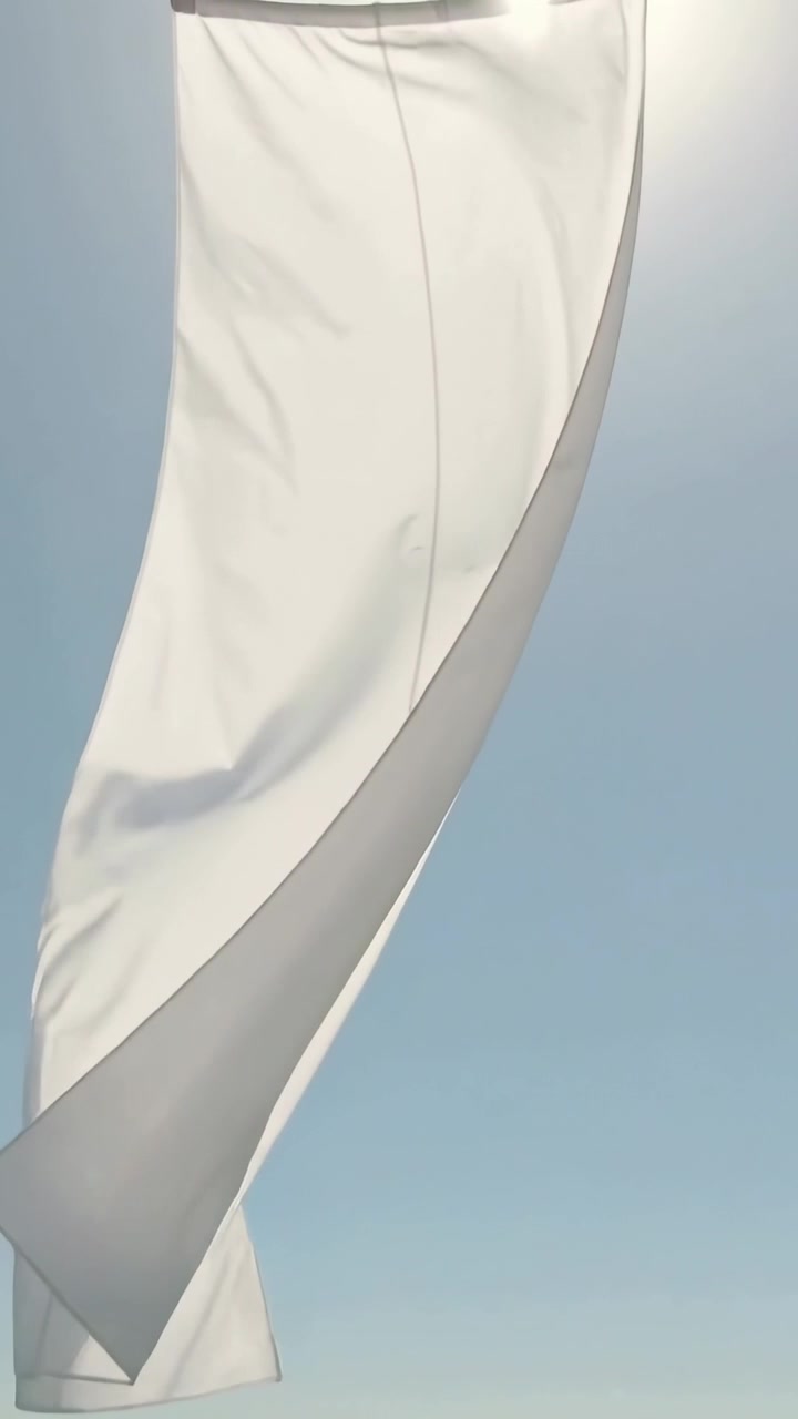 Video vertical: Tela blanca retorciéndose y desplegándose contra el cielo azul claro por el viento, con costuras iluminadas por el sol.