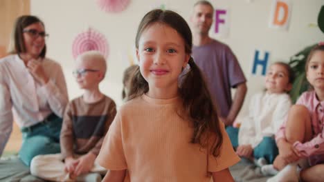 una niña feliz con ojos azules en una camiseta naranja mira a la cámara y sonríe contra el fondo de su primera lección con los maestros en preparación para la escuela. niña preescolar preparándose para la escuela con sus maestros y un grupo de niños