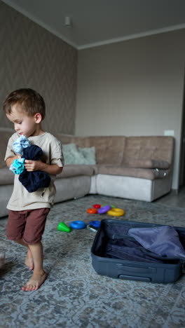 little cute kid picks his clothes from the floor and suitcase every time dropping one. energetic toddler tosses his things up. vertical video.