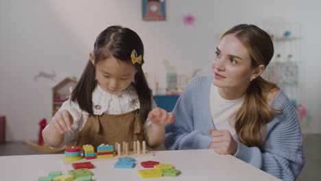 Asiatisches-Kleines-Mädchen-In-Einer-Montessori-Schule,-Das-Mit-Formen-Spielt,-Die-Sich-Stapeln,-Während-Ein-Kaukasischer-Lehrer-Ihr-Hilft