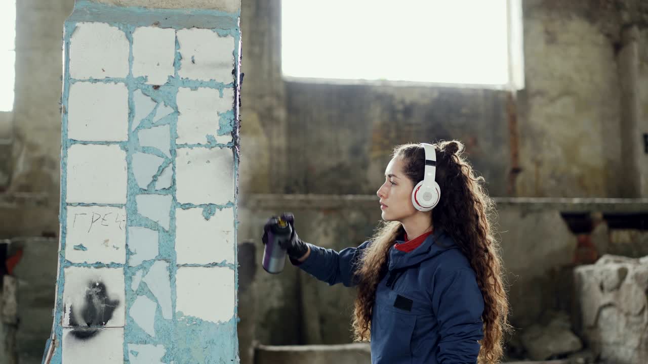una chica atractiva se concentra en decorar una vieja columna sucia con graffiti en un edificio abandonado usando pintura de aerosol. una joven está escuchando música con auriculares.