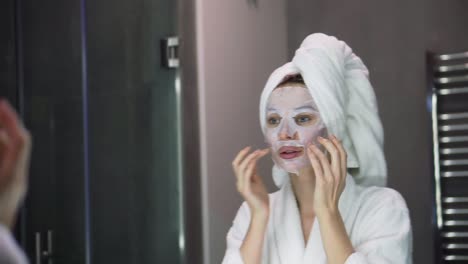 portrait of a woman in bathrobe with white mask for moisturizing on face, looking at the mirror