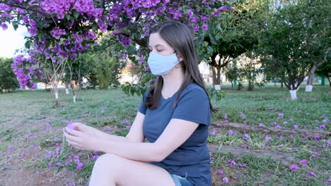 caucasian woman with face mask sitting in park collecting pink flower from spring tree