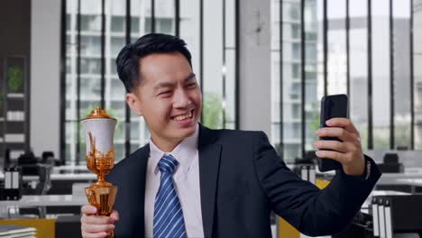 close up of asian business man in a suit and tie with a gold medal holding a gold trophy and taking photo on smartphone as the first winner in office