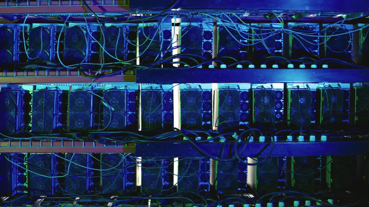 Multiple rows of Bitcoin mining rigs with cooling fans and tangled cables, lit by blue light in a data center. Pan right, close up