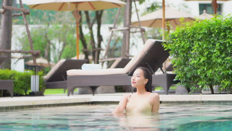 woman with wet hair inside swimming pool water with lounge deckchairs and parasols behind in tropical hotel - slow motion