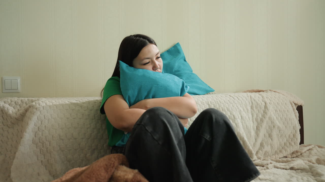 침대에 앉아 베개와 곰인형을 들고 있는 여성, 담요가 덮여 있고 은은한 조명이 아늑하고 친밀한 분위기를 연출하며, 차분한 작가가 사색에 잠겨 휴식을 취하고 있고, 가정적인 침실의 디테일이 눈에 띄어요.