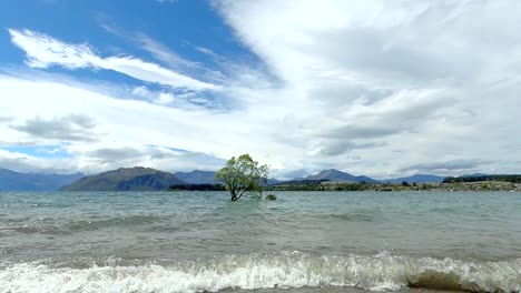 A-peacful-scene-of-a-lone-tree-in-the-middle-of-a-large-lake-with-mountains-in-the-background-and-blue-cloudy-sky