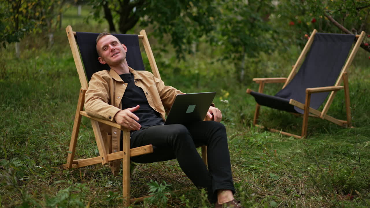 Happy relaxed man laid back in the garden chair. Male freelancer working remotely in the autumn garden. Man speaking on video chat on his laptop.