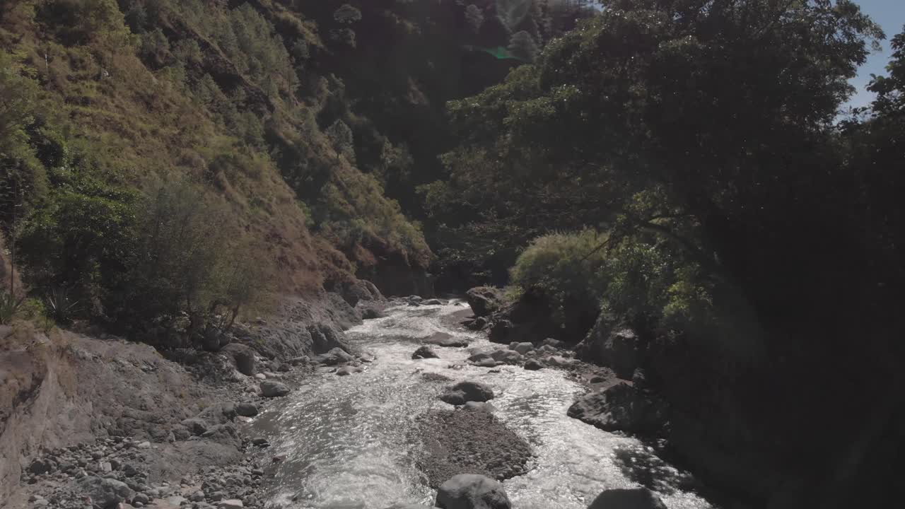 Metal wire suspension bridge spanning over river in remote wilderness terrain aerial flying under through following river approaching