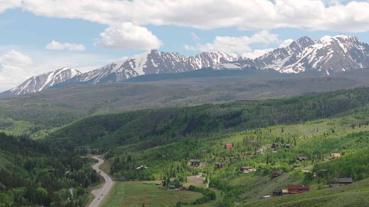 vista aérea de cerca a la derecha de la hermosa cordillera de colorado con picos cubiertos de nieve en un día soleado de cielo azul en el verano con campos verdes, árboles y casas de montaña