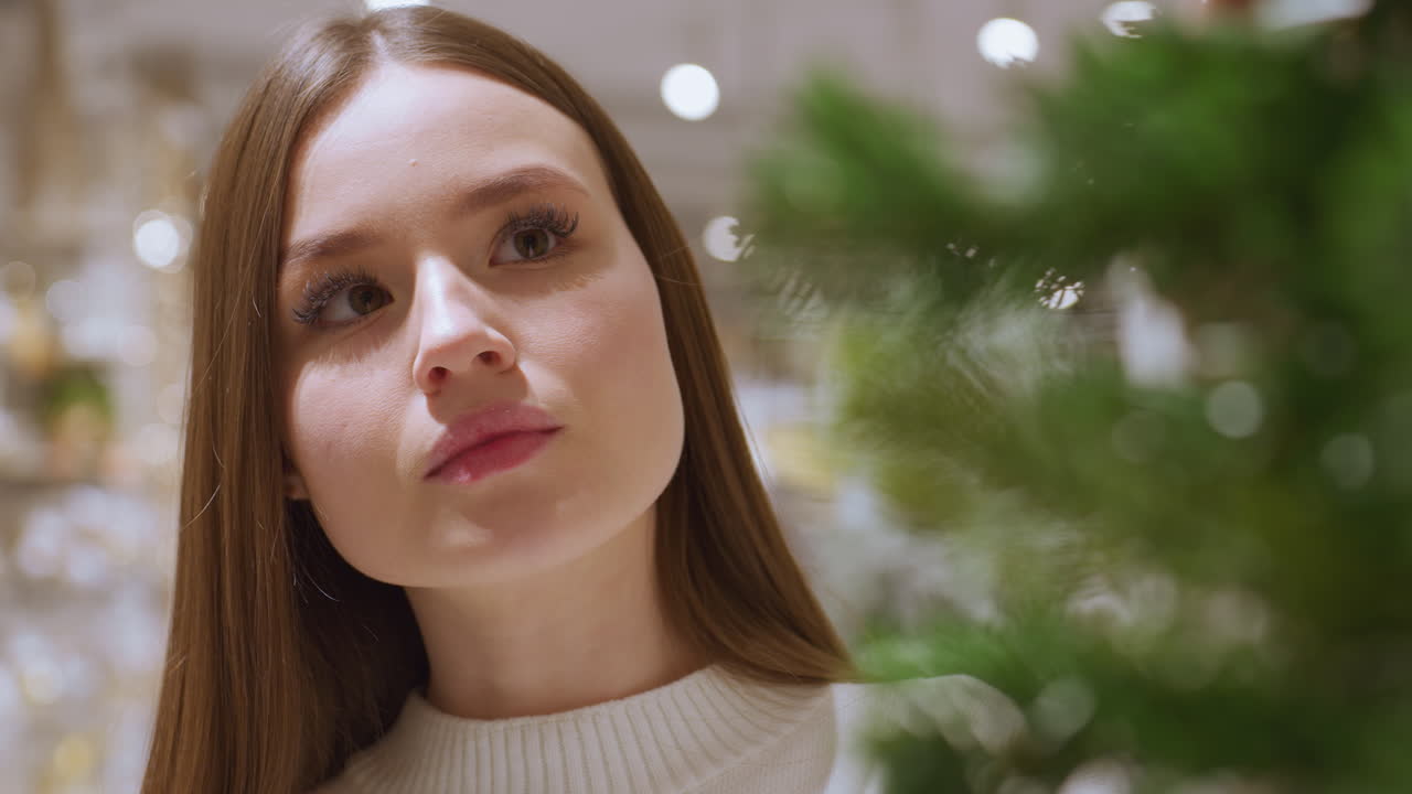 Close-up of young woman admiring beautifully hanging tinsel in a festive store, she is surrounded by holiday decorations, including garlands and shimmering lights in a seasonal shopping mall