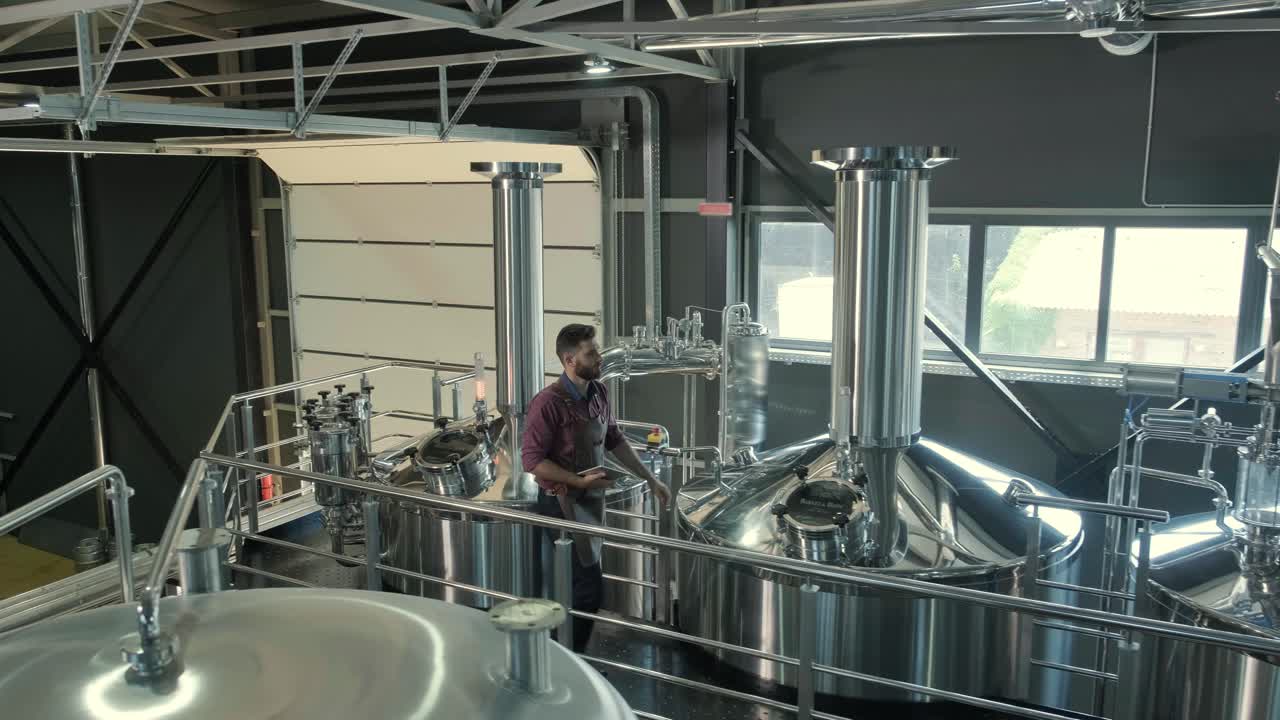 Young male brewer wearing a leather apron supervise the process of beer fermentation at a modern brewery factory