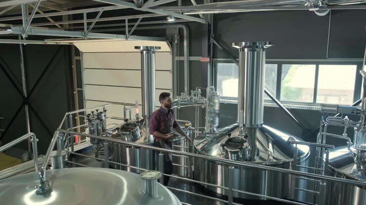 Young male brewer wearing a leather apron supervise the process of beer fermentation at a modern brewery factory