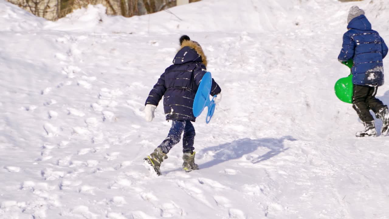 Children having fun, playing and laughing on snowy winter walk in nature. Frost winter season. Cute kids with plastic sledges walk up a hill outdoors on winter day. Winter games and fun