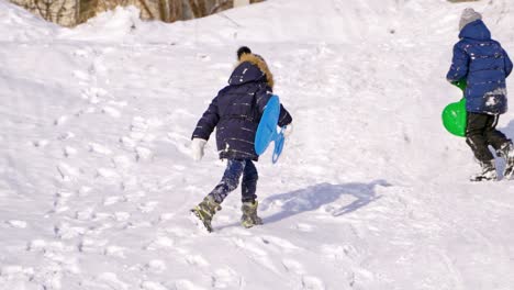 children having fun, playing and laughing on snowy winter walk in nature. frost winter season. cute kids with plastic sledges walk up a hill outdoors on winter day. winter games and fun