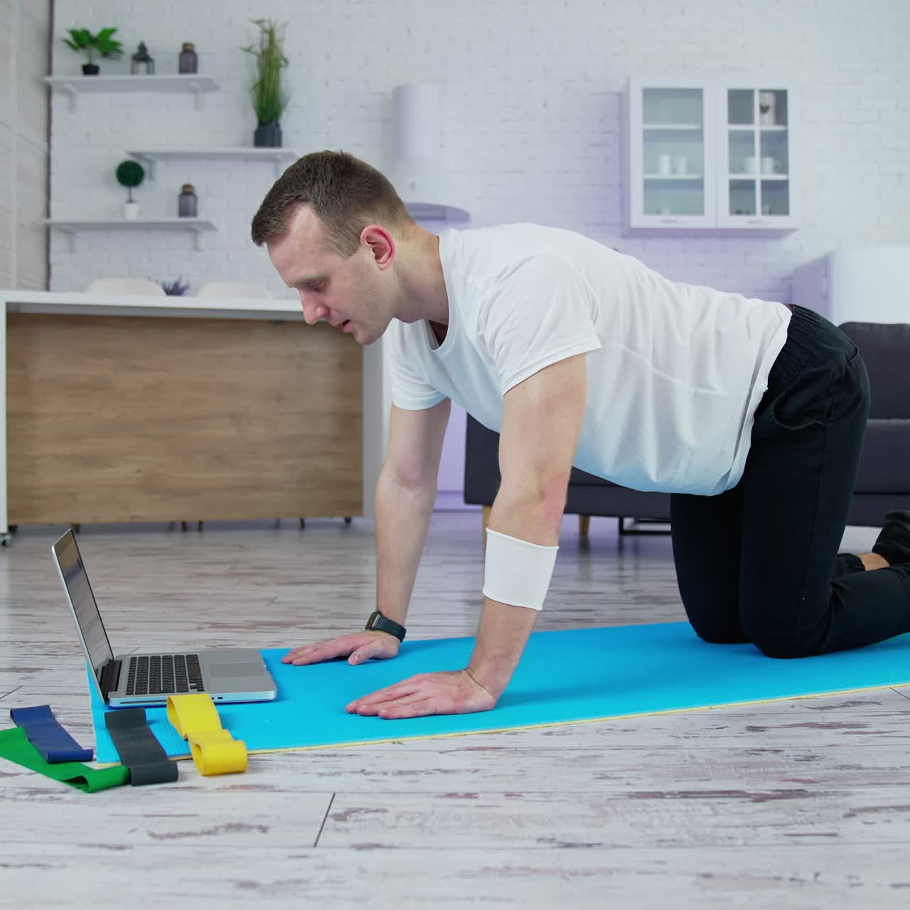 Guy in white t-shirt and black pants training in the kitchen. Young man doing exercise on a mat and looking into a laptop. Slow motion