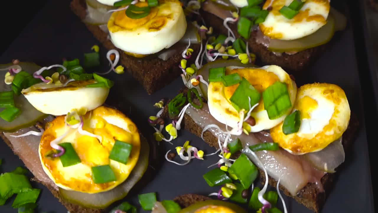 Vue de dessus en gros plan de sandwiches traditionnels baltes aux sprats sur pain de seigle, avec des œufs durs tranchés, des cornichons, des pousses et des herbes fraîches, le tout disposé sur une assiette sombre. Servi comme collation festive estonienne, une tradition culinaire pour les fêtes.