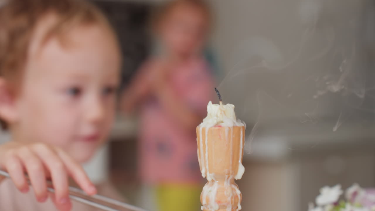 Focused child carefully uses snuffer to extinguish burning candle in calm indoor setting while another child watches from background, demonstrating safety learning and responsibility