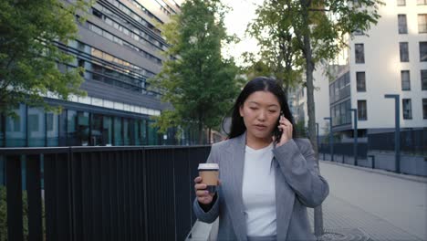 Geschäftschinesin-Telefoniert-Und-Hält-Eine-Tasse-Kaffee-In-Der-Hand,-Während-Sie-Durch-Die-Straße-Geht.