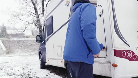 una persona con una chaqueta azul abre la puerta trasera de una caravana blanca estacionada en una carretera nevada cerca de edificios residenciales