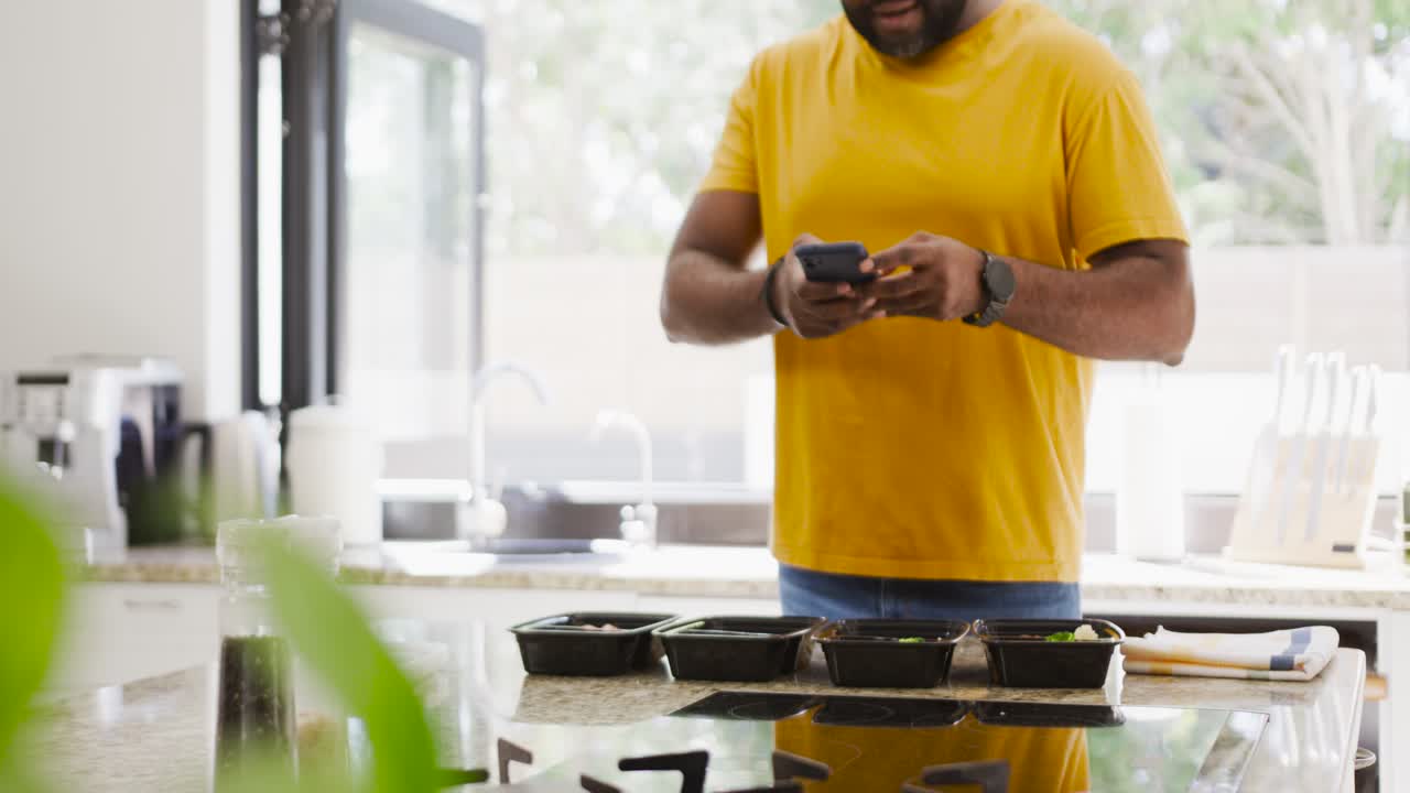 African American man in yellow tee seeing notification, tapping phone at island, sharing four trays