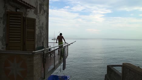 Man-ponders-on-balcony-of-Mediterranean-home-over-ocean,-birds-fly-past,-slow-motion