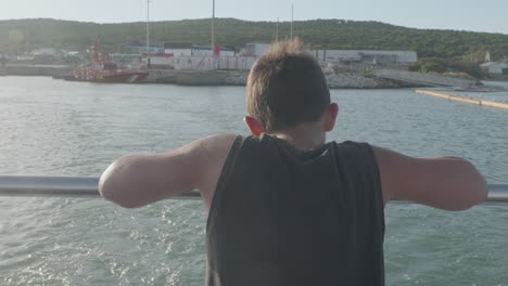 niño apoyado en la popa de un barco que se aleja del puerto