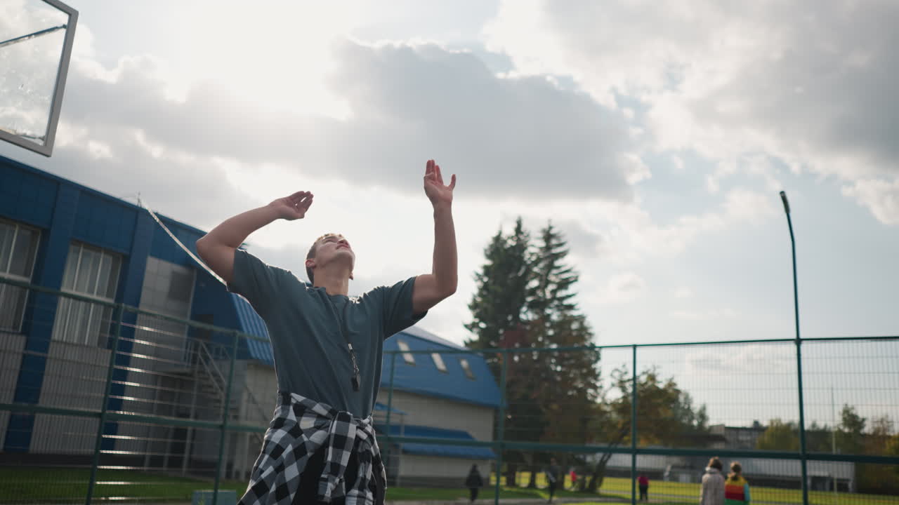 joven con camisa alrededor de la cintura salta para golpear el voleibol en la arena deportiva con el fondo de personas caminando, cancha al aire libre y equipos deportivos visibles