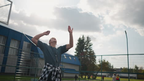 youngster with shirt around waist jumps up to slam volleyball in sports arena with background of people walking, outdoor court and sports equipment visible