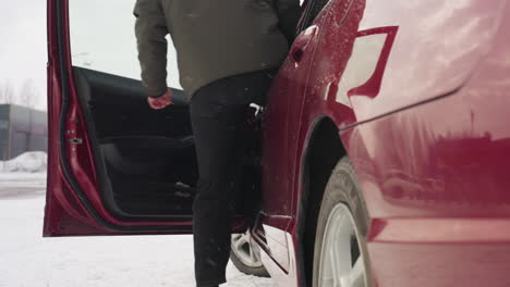 winter day man in winter jacket opens red car door and enters vehicle against snowy industrial backdrop, water droplets on glossy surface reflect nearby buildings and parked cars during cold season