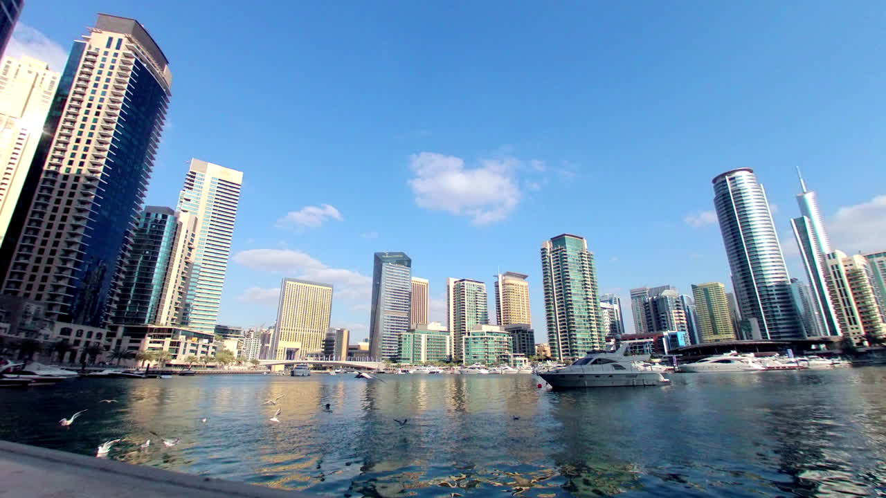 Skyscrapers rise above the marina in a wide fisheye view with boats and open sky