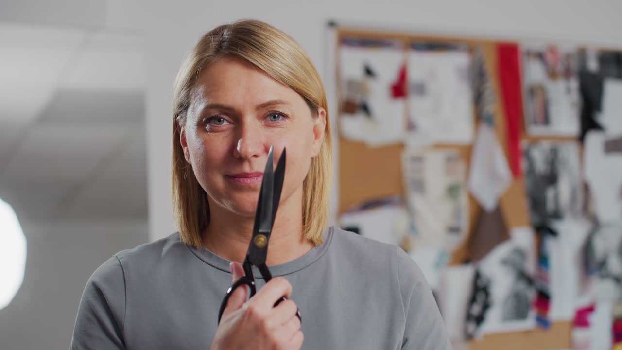 Fashion designer lifting black scissors across face pretending to cut material while smiling joyfully, standing inside bright sewing workspace decorated with creative boards and colorful textiles