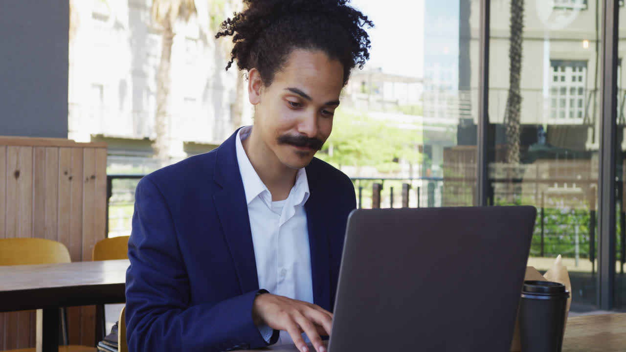 Mixed race man with moustache sitting at table outside cafe using laptop