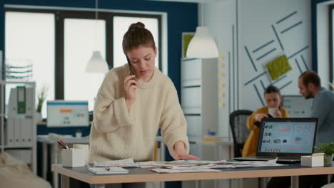 busy woman in startup office talking on smartphone looking at papers with sales charts