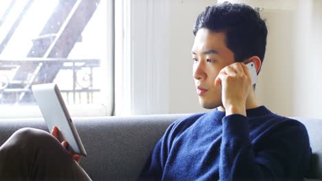 man talking on mobile phone while using digital tablet in living room 4k