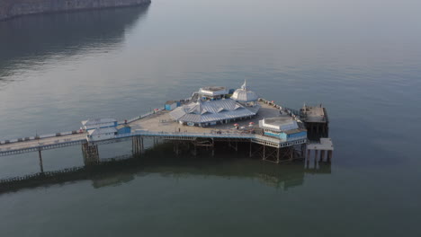 Aerial-view-of-the-end-of-Llandudno-pier-flying-left-to-right-around-it-as-the-sun-begins-to-set,-Llandudno,-North-Wales,-UK