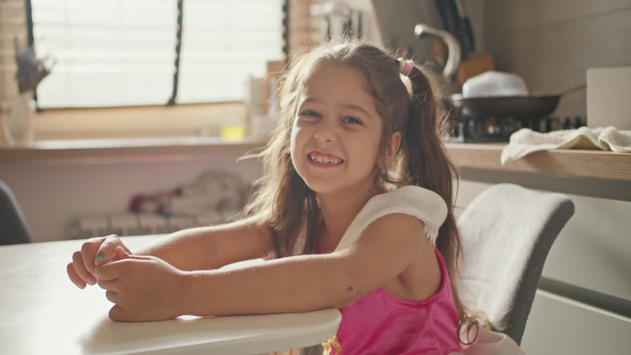retrato de una feliz niña morena en un vestido rosa que se sienta en la mesa de la cocina en una cocina moderna durante el día. una niña con un vestido de hada se sienta a la mesa en la cocina, se regocija y se divierte
