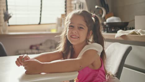 Portrait-of-a-happy-little-brunette-girl-in-a-pink-dress-who-sits-at-the-kitchen-table-in-a-modern-kitchen-during-the-day.-A-little-girl-in-a-fairy-dress-sits-at-the-table-in-the-kitchen,-rejoices-and-has-fun