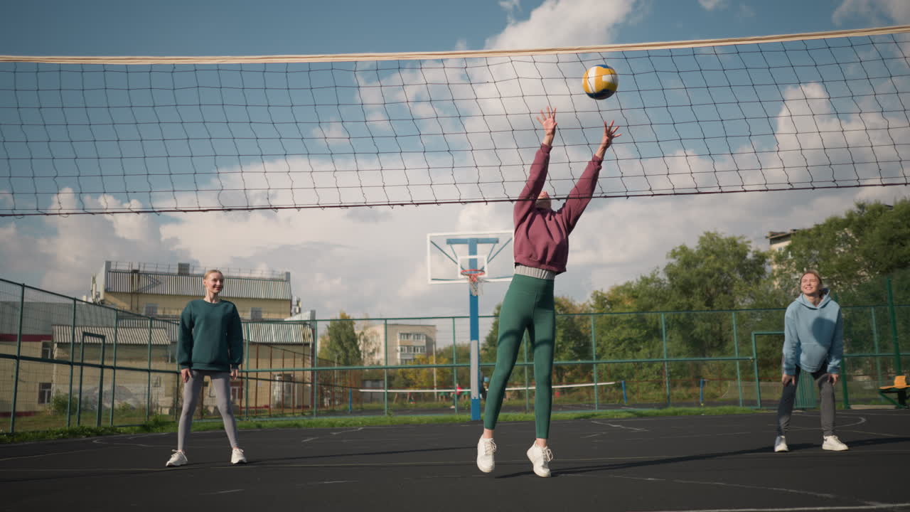중앙에 있는 여자와 함께 코트에서 서 있는 배구 초보자 들 이 공 을 기 위해 준비 하고 있으며, 다른 선수 들 이 지켜보고 있으며, 초록색 과 건물 이 배경 에 있으며, 야외 스포츠 환경 이 있다.