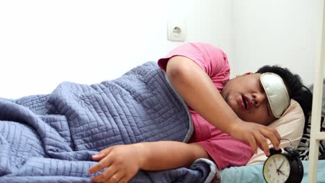 sleepy man wearing eye mask yawning while lying in bed with alarm clock at his side