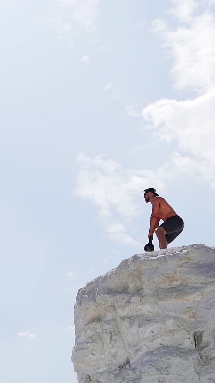 View from below on a healthy man training with heavy weight on a hill. Bodybuilder lifting kettlebell on the natural sky background. Shirtless man during workout outdoors. Vertical video