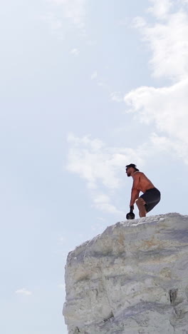 view from below on a healthy man training with heavy weight on a hill. bodybuilder lifting kettlebell on the natural sky background. shirtless man during workout outdoors. vertical video