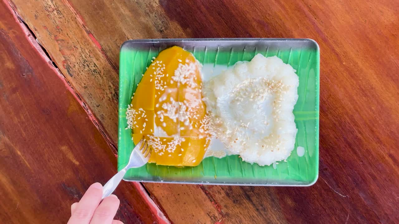 A person enjoys mango sticky rice at a clifftop restaurant in Phuket, Thailand. Bright lighting highlights the vibrant colors