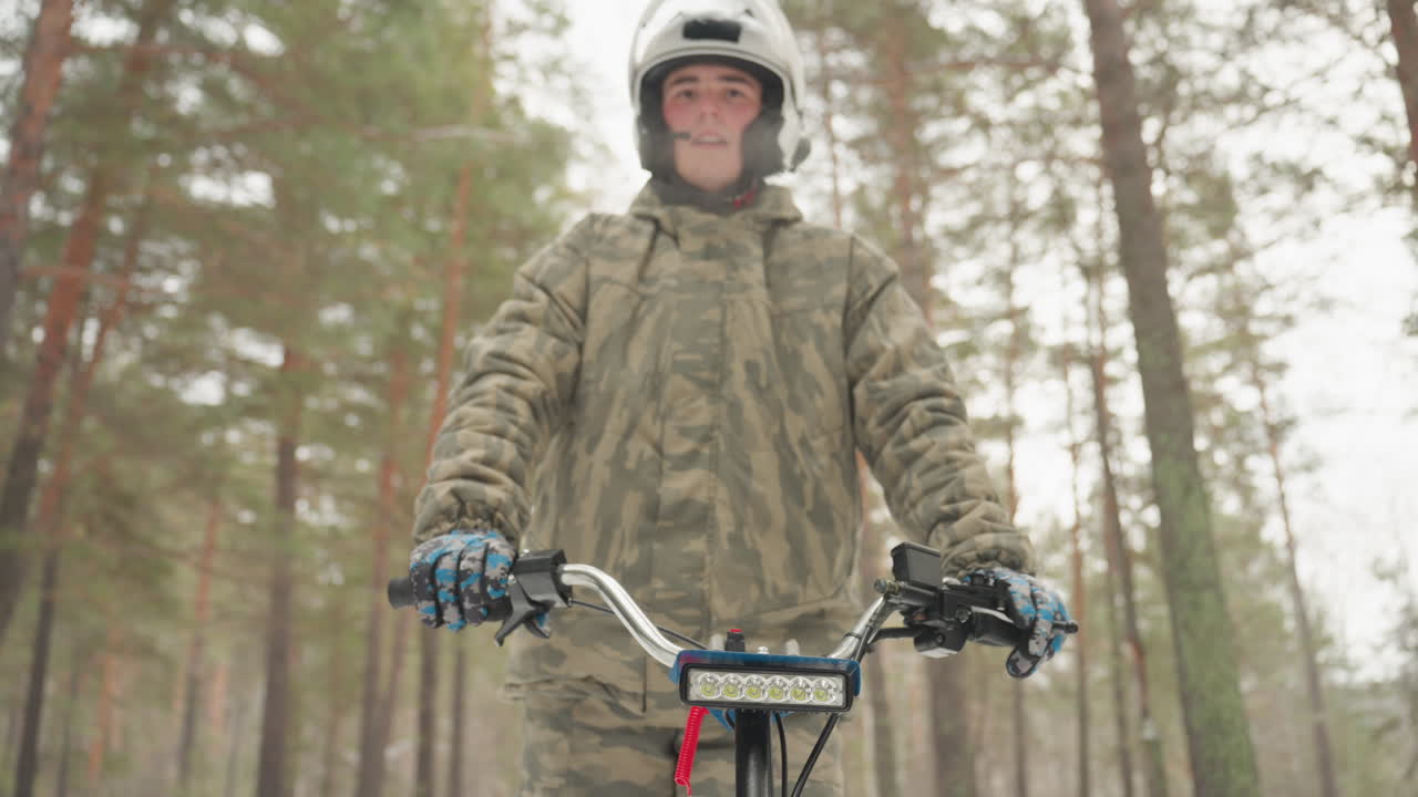 Rider on atv snowy forest. Camouflage jacket and blue gloves grip chrome handlebars, helmeted rider ready for extreme trail run, pine trunks and soft snow create cold, focused atmosphere, closeup