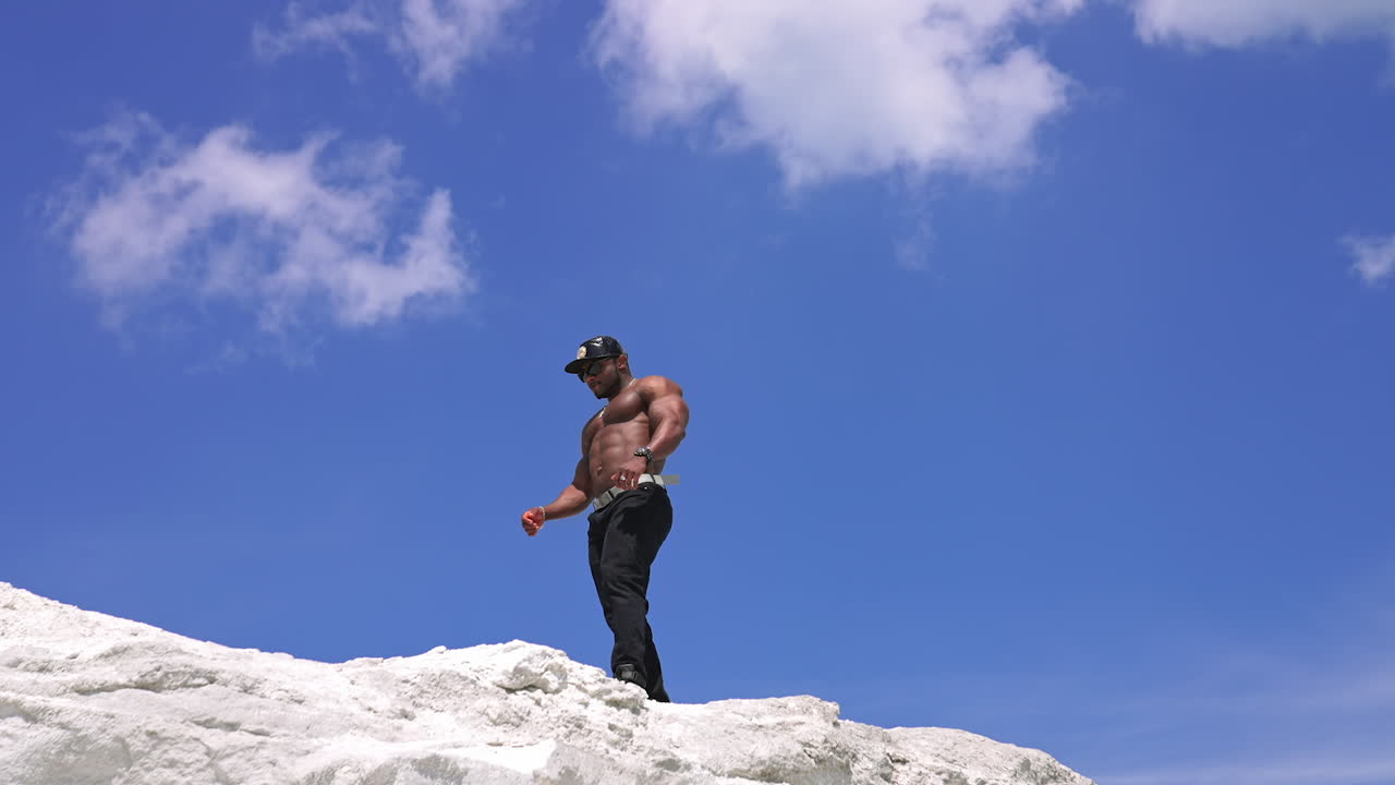 Afro american athlete on blue sky background. Dark skinned bodybuilder without shirt with muscular body walking along white hill at sunlight. View from below.