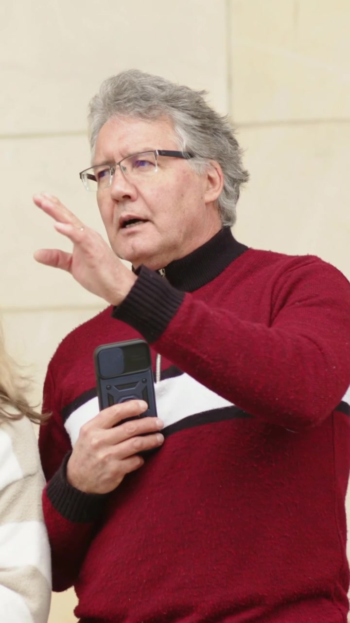 Video verticale: Uomo con occhiali e maglione rosso alza la mano, parla vicino a un muro di pietra e mostra il telefono.