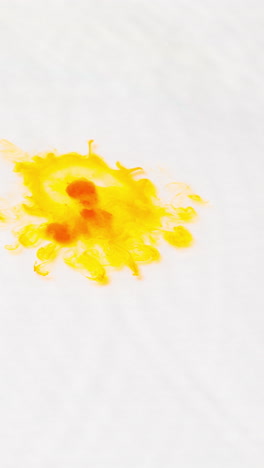 tomada de video vertical aérea de una gota de pintura o tinte amarillo caída en el agua contra un fondo blanco para crear un patrón de colores giratorios