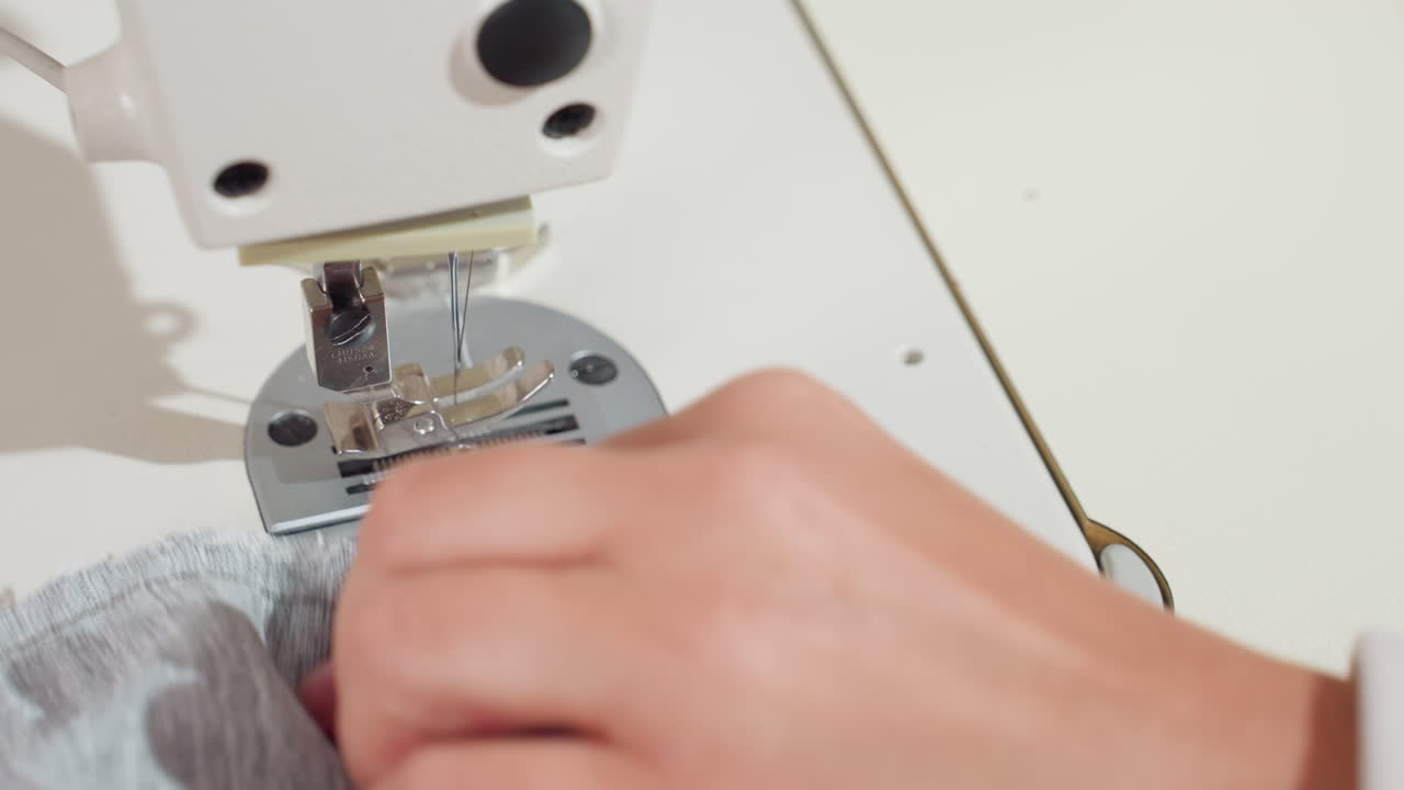 Close up of human hand guiding light gray fabric under sewing machine needle during stitching process, showing focus and detail in textile construction inside bright workshop environment