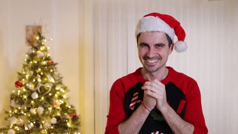 un hombre con sombrero de santa se emociona mientras se frota las manos en la sala de estar de una casa moderna decorada con un árbol de navidad para las vacaciones navideñas de invierno